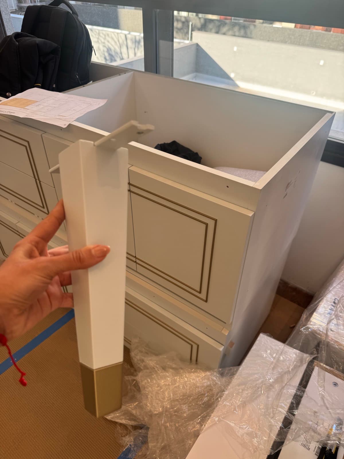 A hand holding a white furniture leg with a brass foot, in front of a fitted drawer unit